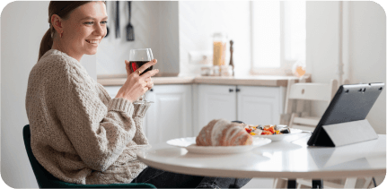 A young girl is having a breakfast with a glass of wine in her hand and looking at the tablet smilingly.